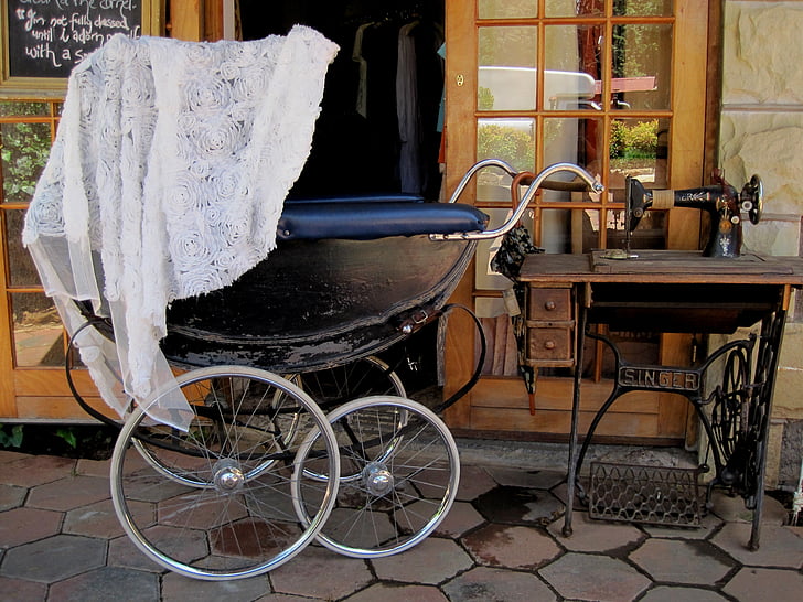 black perambulator, large wheels, old fashioned, old sewing machine, white lace covering, grey floor paving, shop entrance